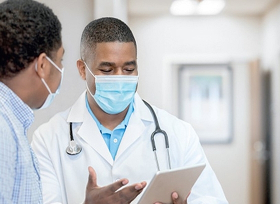 Doctor wearing a mask and a white coat reviewing a tablet with a patient in an exam room