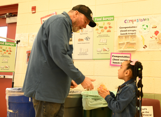 adult helping child sort cafeteria food waste at collection station