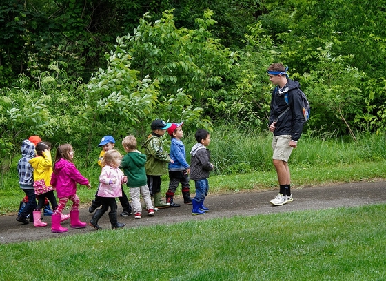 An activity leader guiding a group of toddlers along a park path.