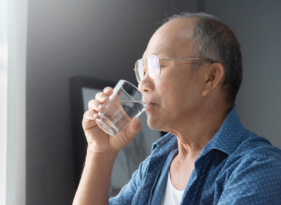 A senior asian man drinking a glass of water while looking out the window.
