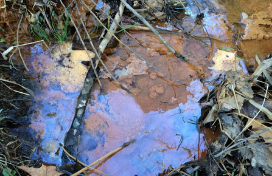 A broken, orangish, oily sheen of iron floc sits in a puddle of water. 