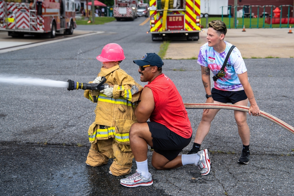 Camper in turnout gear uses fire hose with assistance of firefighter.